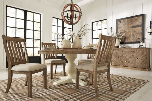 view of a modern farmhouse style dining room with an oval light wooden table, four chairs around it, and a matching natural wooden sideboard behind it. The room has white walls and a large wooden accent art piece above the sideboard hanging on white panel walls with large rustic windows.