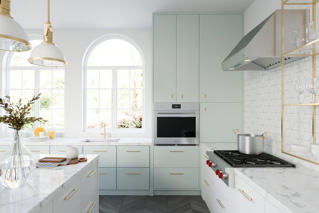 Front view of a modern kitchen with a Wolf E series wall oven built into light blue cabinetry with a Wolf gas rangetop in the white marble counters 