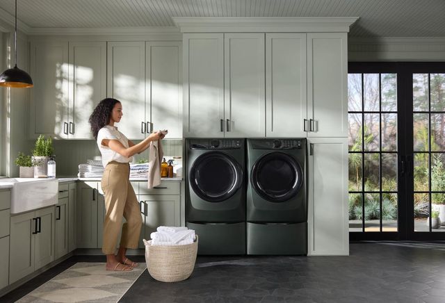 Front view of a modern laundry room with the Electrolux Alpine green washer and dryer as a woman folds clothes in front of them