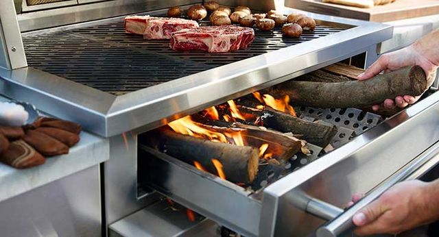 Close view of a Kalamazoo wood fired grill with steaks and potatoes cooking on top, while a person stocks the wood fuel