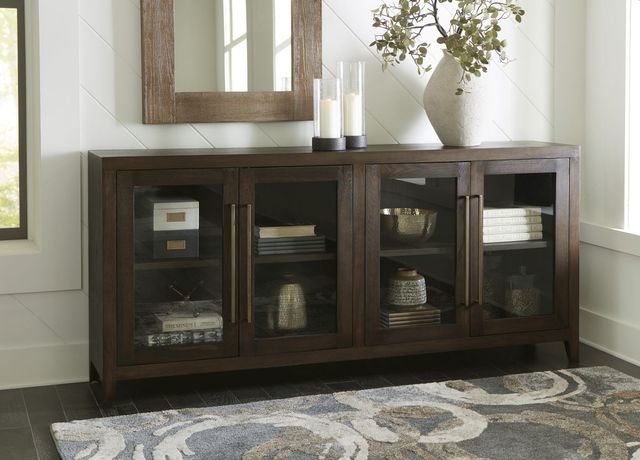front view of a wooden sideboard with glass doors and various decor inside including books and accent vases, with candles on top and a white wall behind it